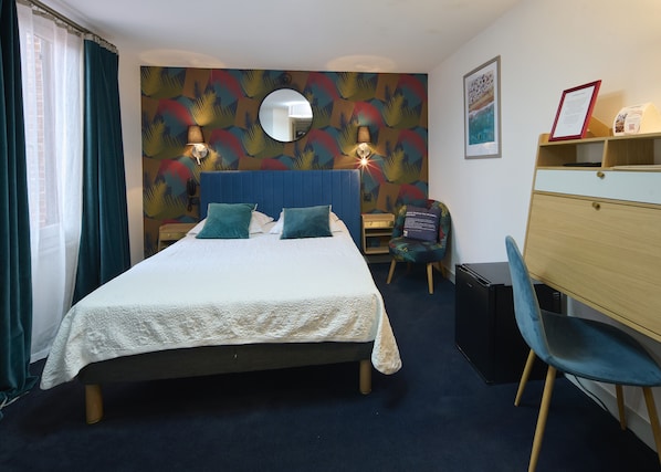 Habitación de hotel con cama, escritorio, silla, lámpara, espejo y pared con estampado de hojas tropicales.