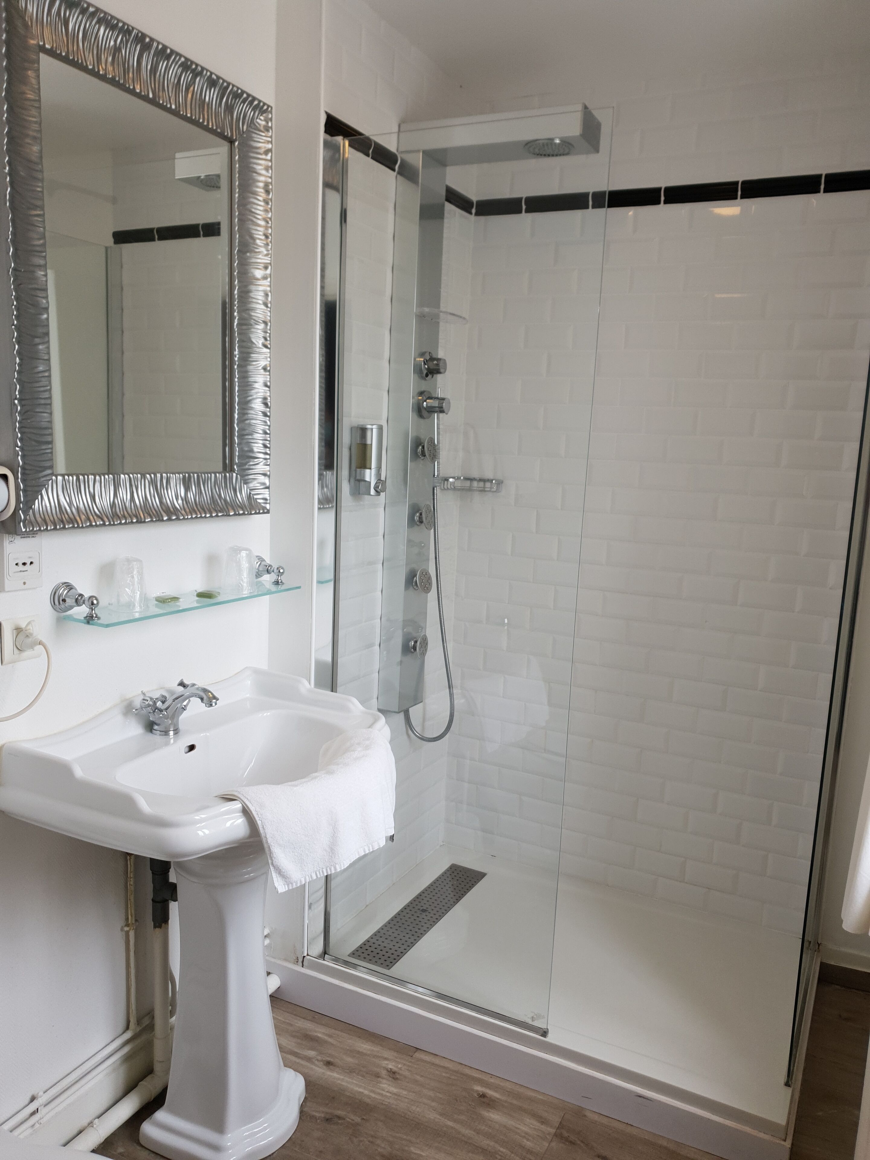 Un baño con lavamanos pedestal blanco, cabina de ducha de vidrio y un espejo grande con marco metálico.