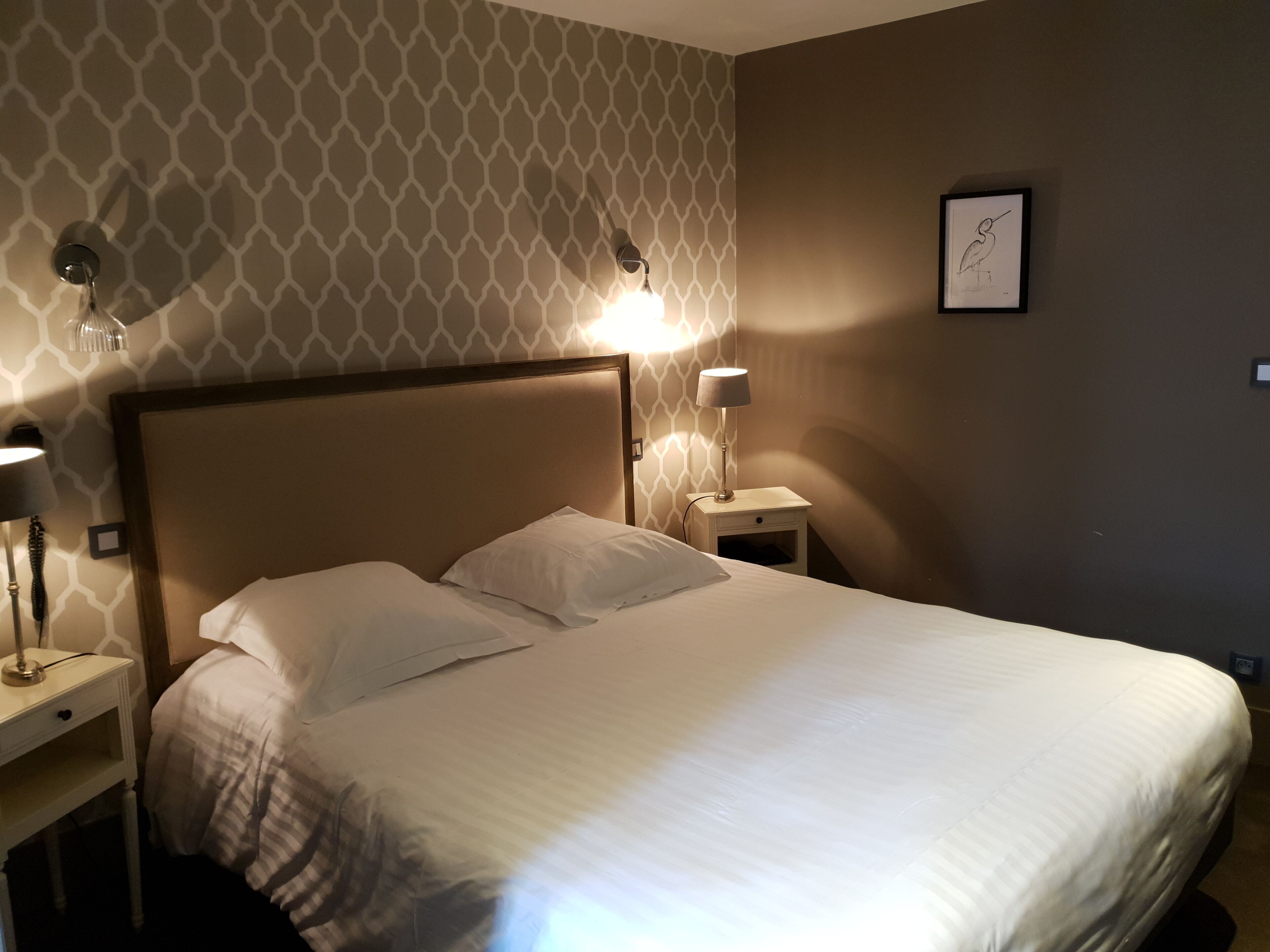 Un lit bien fait, avec du linge de lit blanc, dans une chambre d’hôtel. Il y a deux tables de chevet, chacune avec une lampe et un petit tiroir. Les murs sont recouverts d’un papier peint à motifs géométriques, et un tableau encadré est accroché au mur.