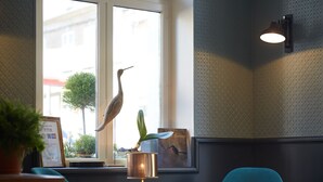 A modern room with teal chairs, a small round table, a lamp, and a window with a view of buildings and trees. - The Originals Boutique, Hôtel La Colonne de Bronze (Saint-Valery-sur-Somme)