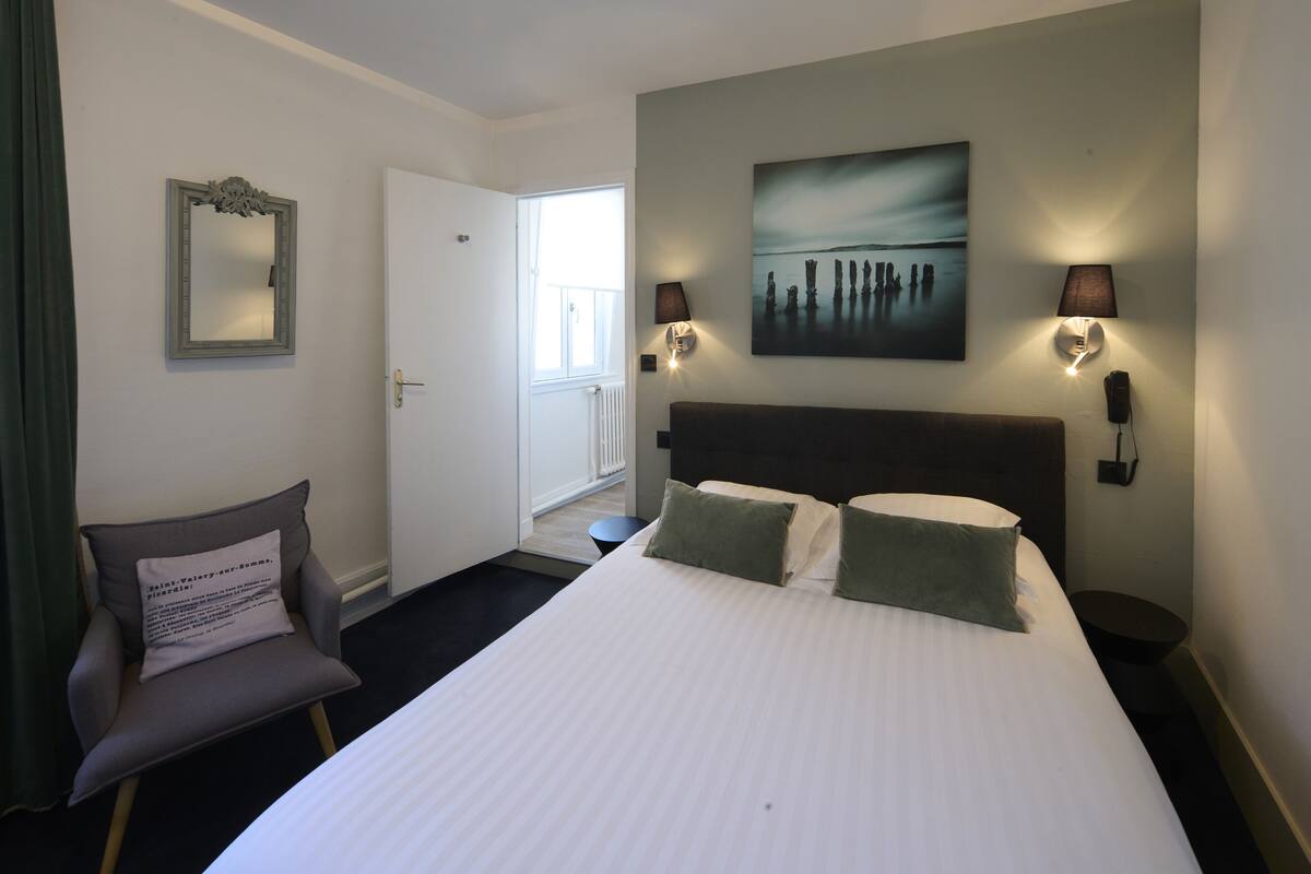 Une chambre d’hôtel avec un grand lit, deux lampes de chevet, une tête de lit, une chaise avec un coussin, un tableau au mur et une porte donnant sur un balcon.