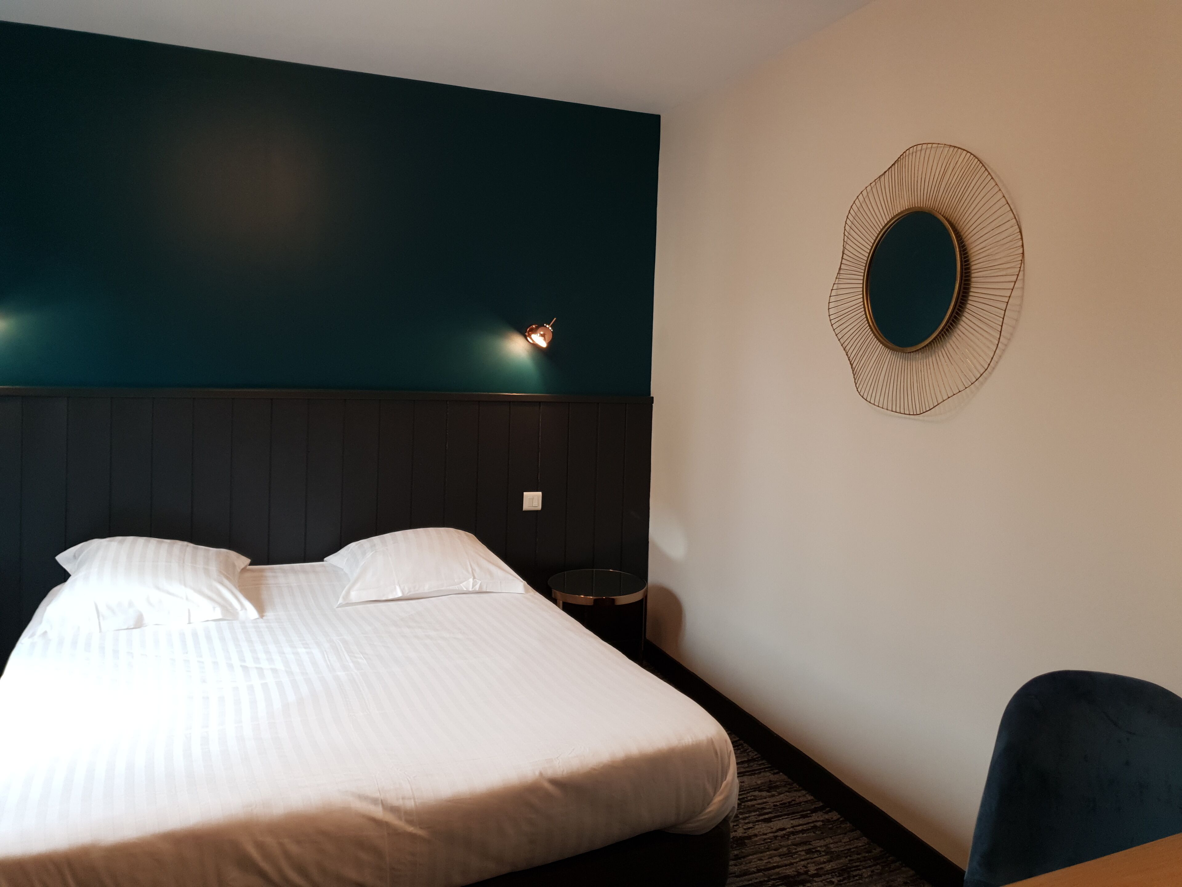 A hotel room with a bed, a nightstand, a chair, and a wall-mounted mirror.