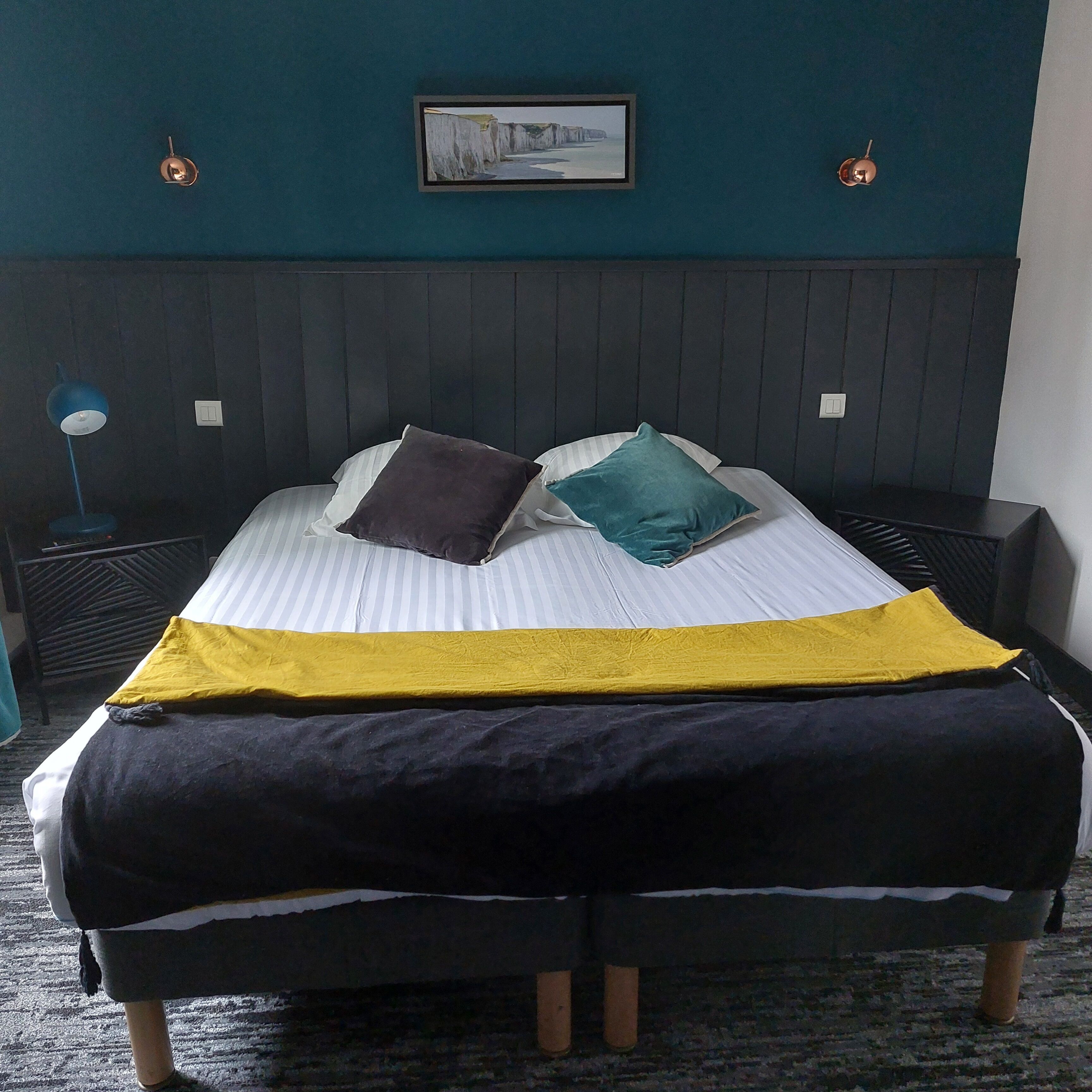 A neatly made bed with striped bedding and two pillows, a dark headboard, a framed picture on the wall, and a blue floor lamp.