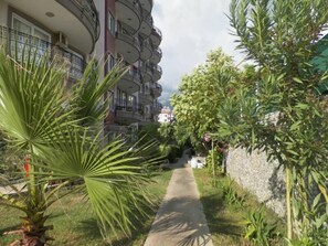 A pathway leading to a multi-story building with balconies, surrounded by greenery and a stone wall.