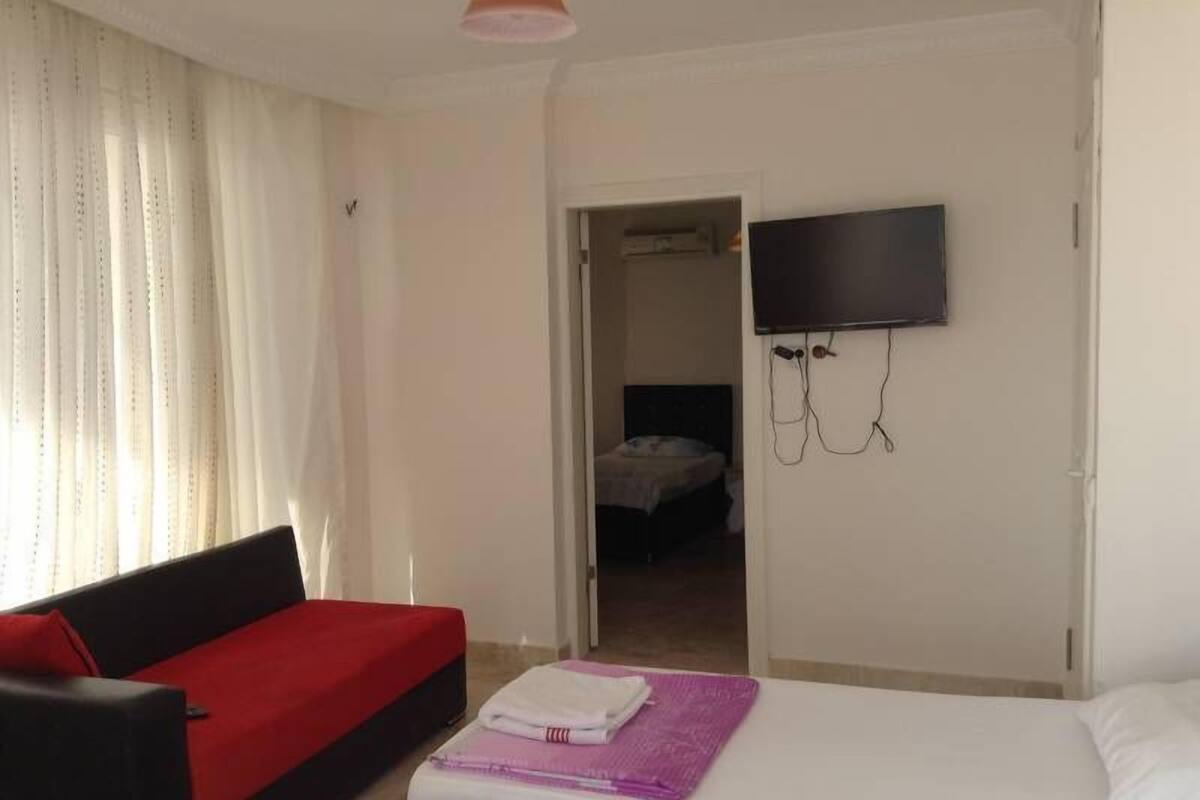 A room with a bed, a red sofa, a TV mounted on the wall, and a door leading to another room.