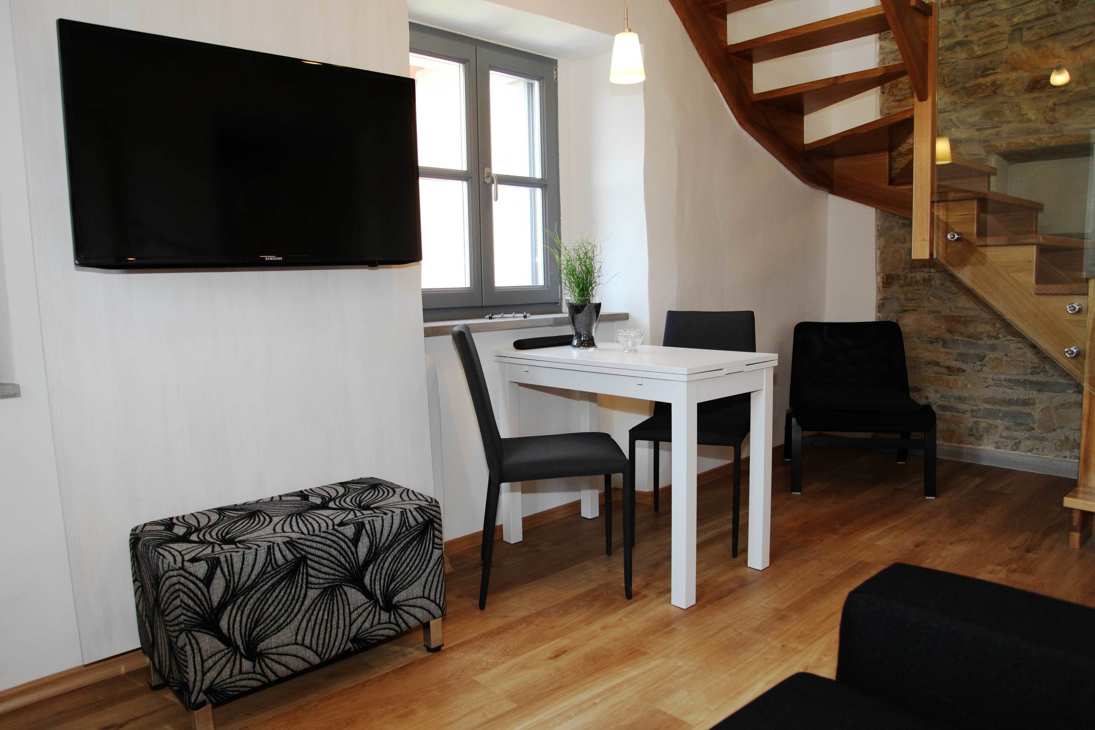 A modern living room with a flat-screen TV mounted on the wall, a dining table with chairs, a bench, and a staircase with wooden railings.