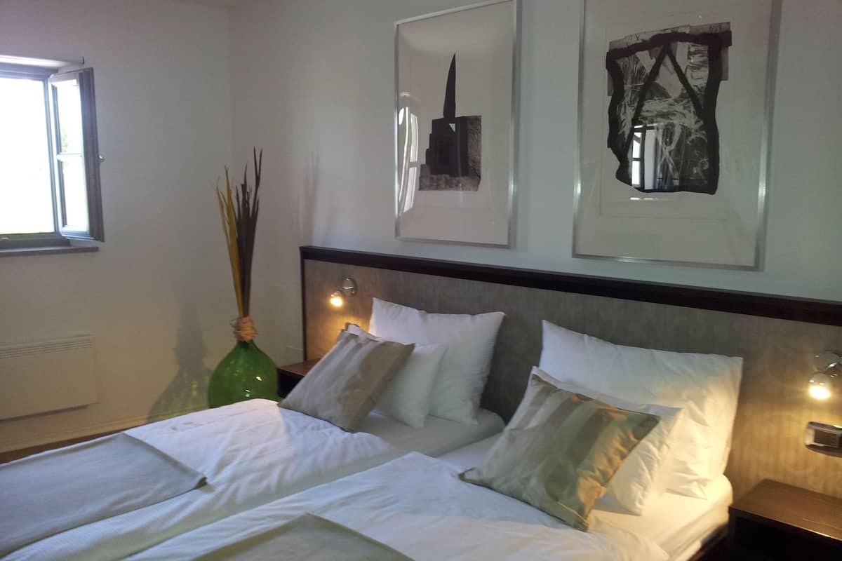 A neatly made bed with white linens and multiple pillows, a bedside table with a lamp, and a window with natural light.