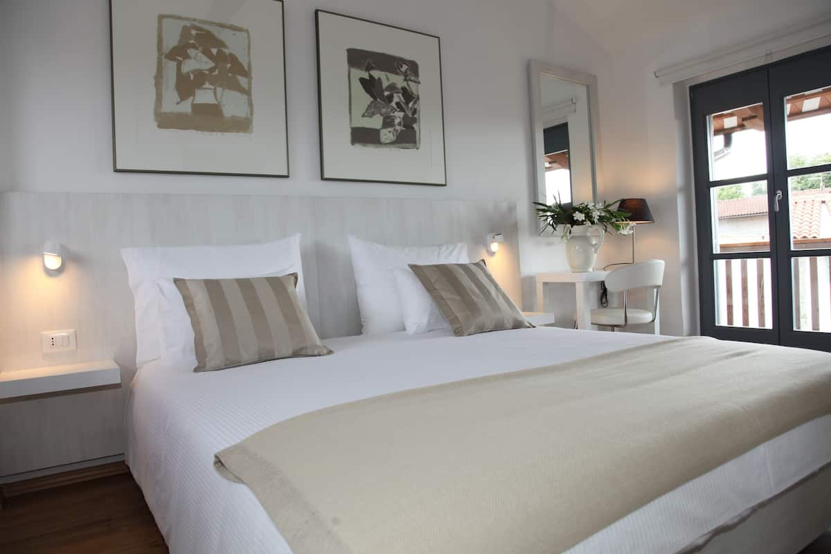 A neatly made bed with white bedding and two pillows, a bedside table with a lamp, and a window with a view of the outside.