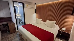 A modern hotel room with a large bed, a wooden headboard, a desk, and a bathroom visible through a glass door. - Hotel León Dorado (Bucaramanga)