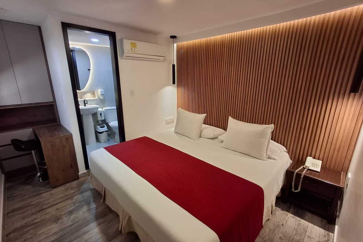 Ein modernes Hotelzimmer mit einem großen Bett, einem hölzernen Kopfteil, einem Schreibtisch und einem durch eine Glastür einsehbaren Badezimmer.