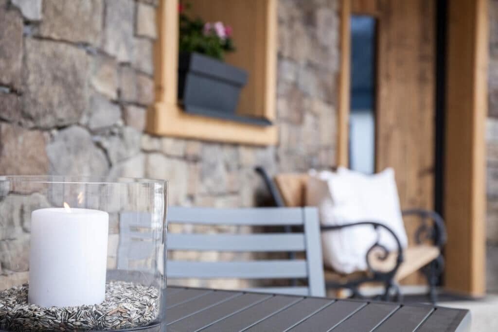 A candle in a glass holder on a table, with a stone wall and a window in the background.