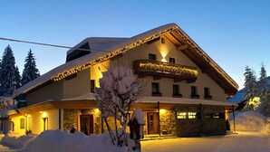 A snow-covered lodge with a sloped roof, stone walls, and a balcony with string lights. - Appartement Ausserladscheider (Leutasch)