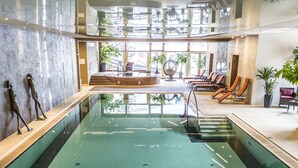 A large indoor swimming pool with a sunken seating area, surrounded by lounge chairs and potted plants. - Hotel OTP Birkenhof (Bad Kleinkirchheim)