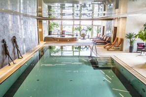 A large indoor swimming pool with a sunken seating area, surrounded by lounge chairs and potted plants. - Hotel OTP Birkenhof (Bad Kleinkirchheim)