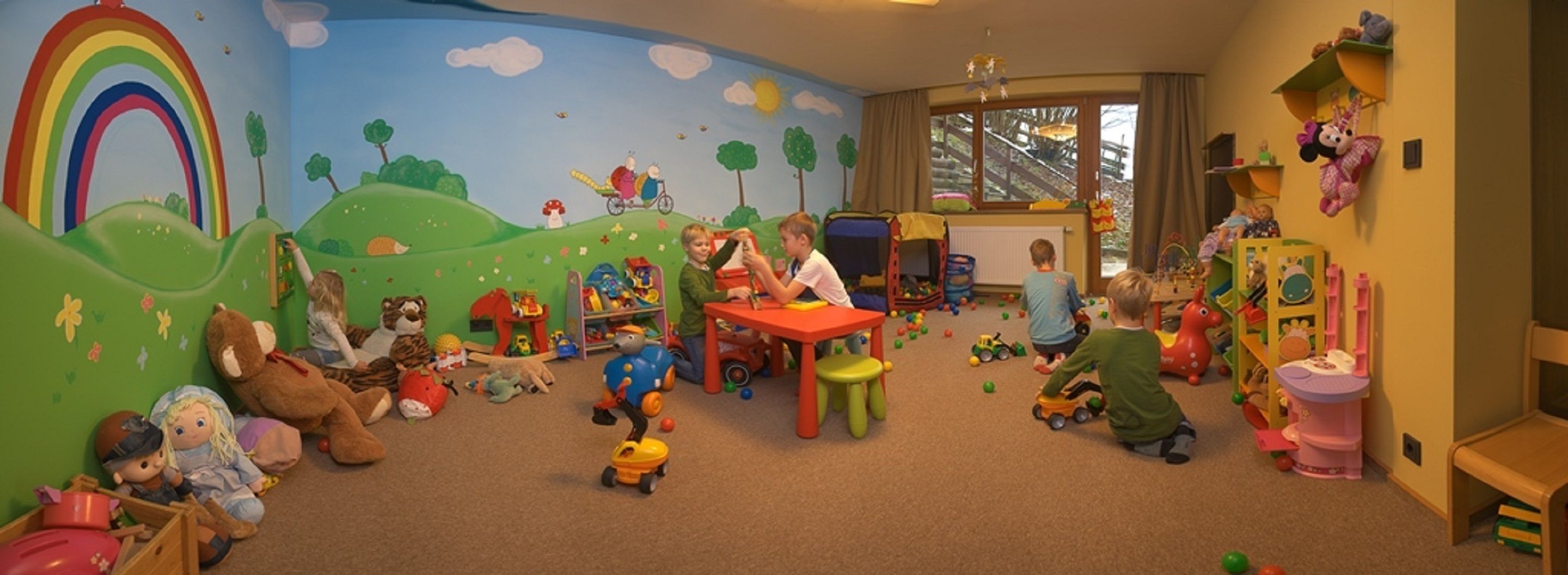 Et legeværelse for børn med et farverigt muralmaleri, diverse legetøj og et bord.