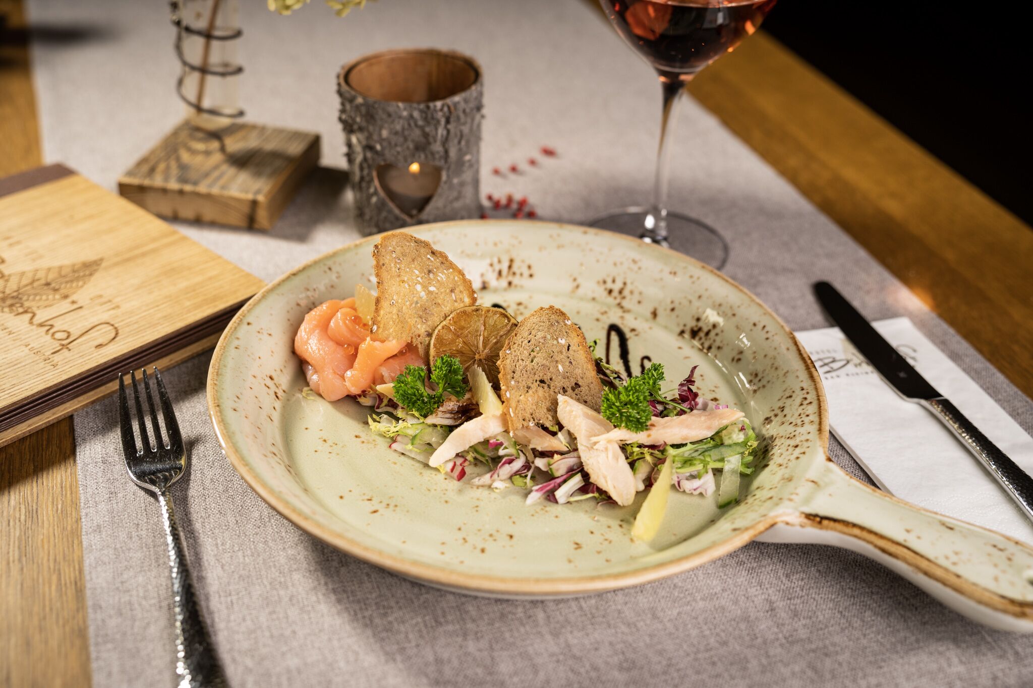 Ein Teller mit Essen, darunter Brot, Lachs und Salat, auf einem Tisch mit Gabel, Messer und Weinglas.