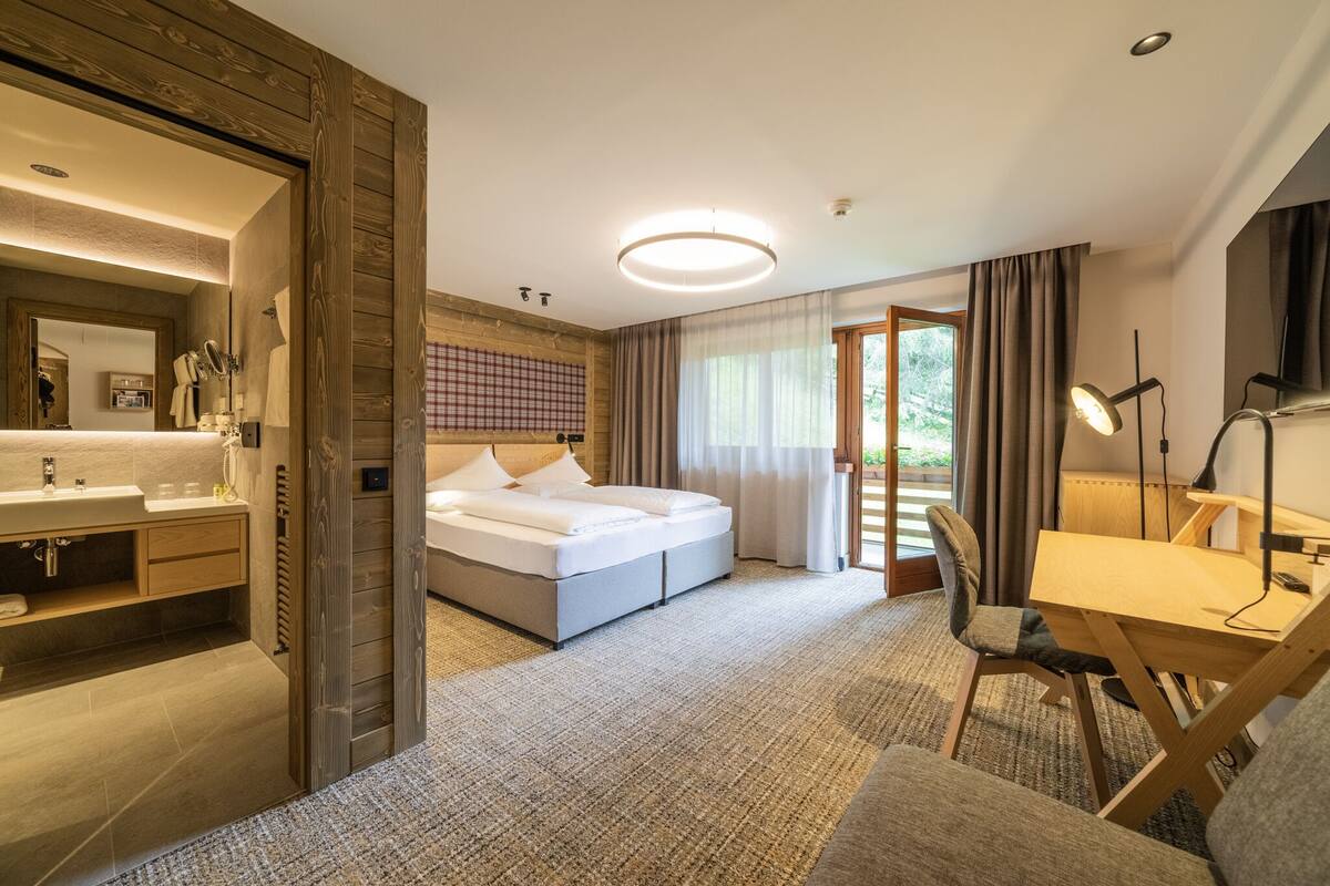 A modern hotel room with a large bed, a desk, a chair, a desk lamp, and a bathroom with a sink and mirror.