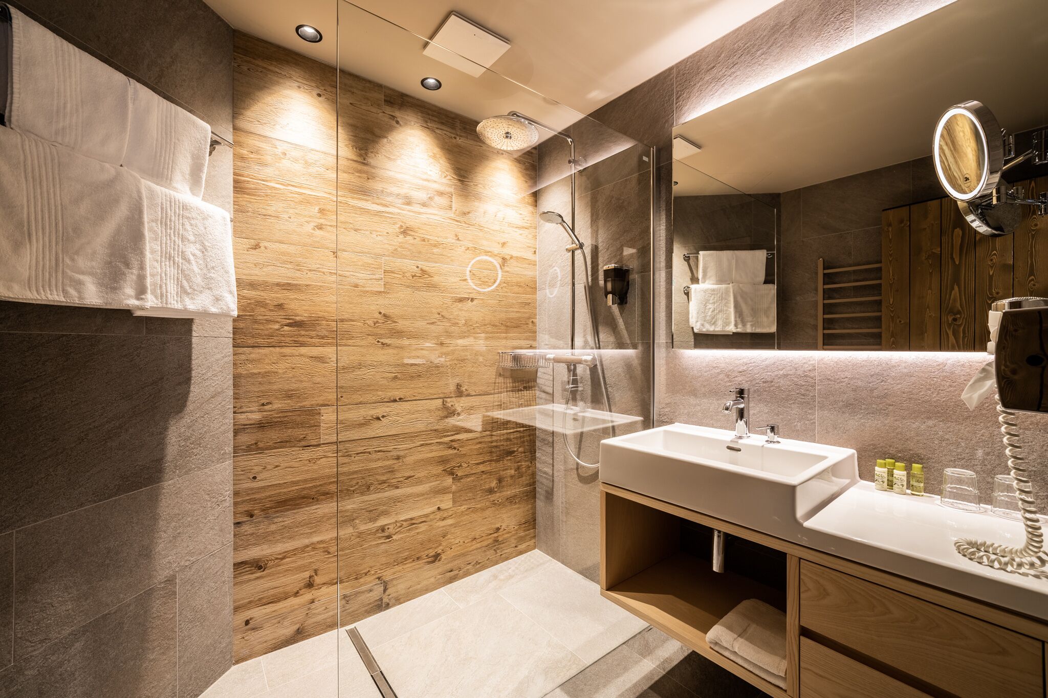 A modern bathroom with a wooden wall, a glass shower enclosure, a white sink, and a mirror with a light.