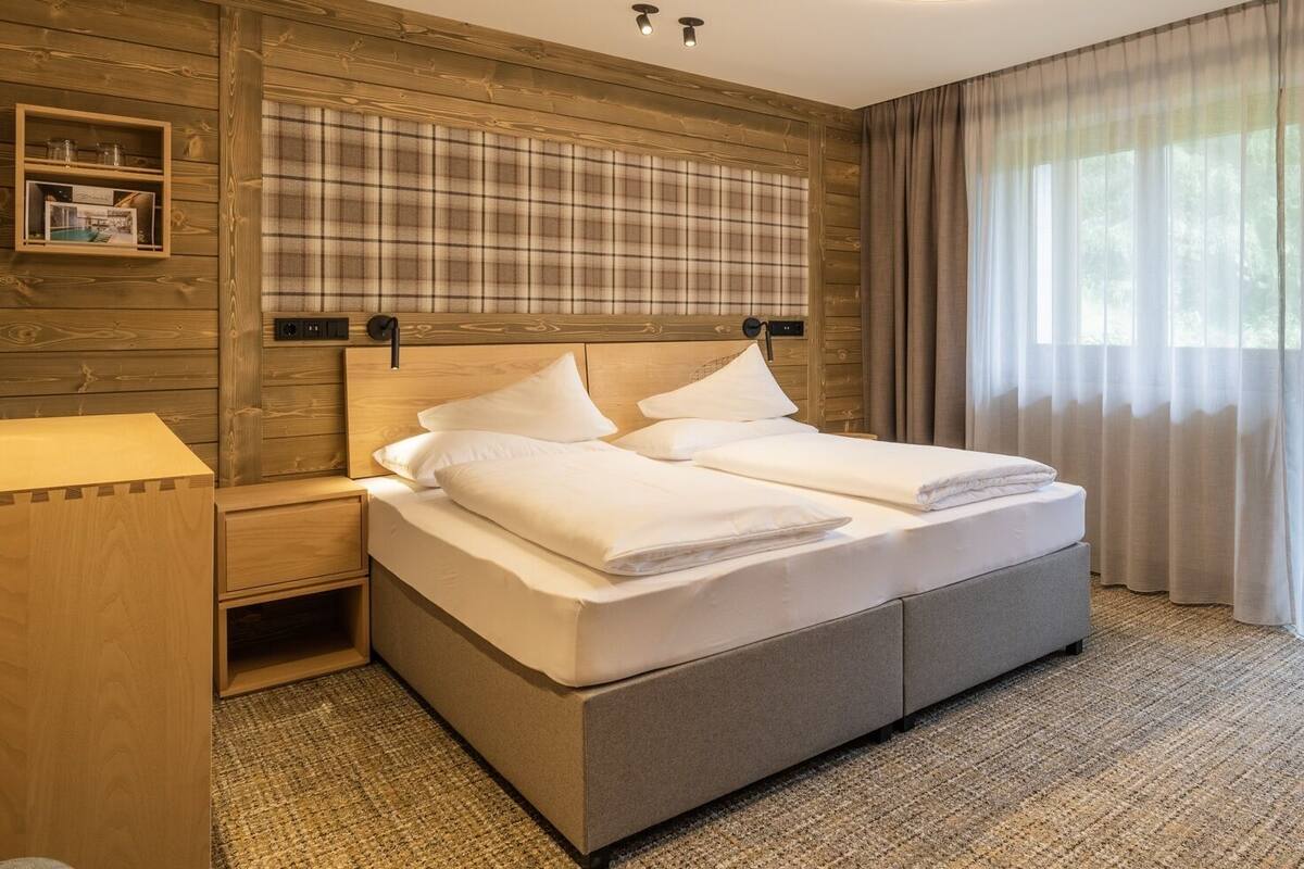 A hotel room with a bed, a bedside table, a headboard with a plaid pattern, and a window with curtains.