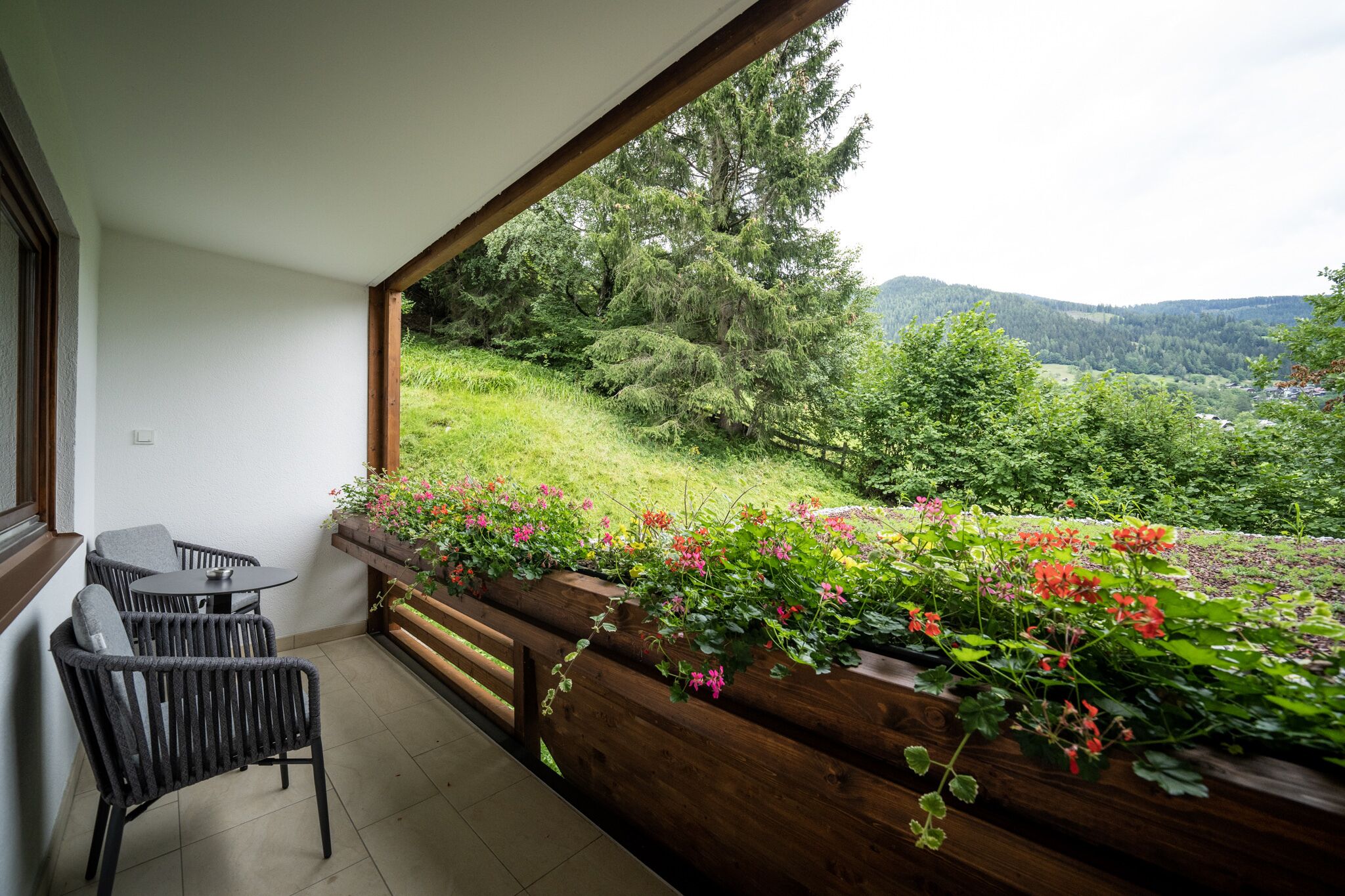 Ein Balkon mit Tisch und Stühlen, Holzgeländer und Blick auf eine üppige grüne Landschaft mit Bäumen und Hügeln.