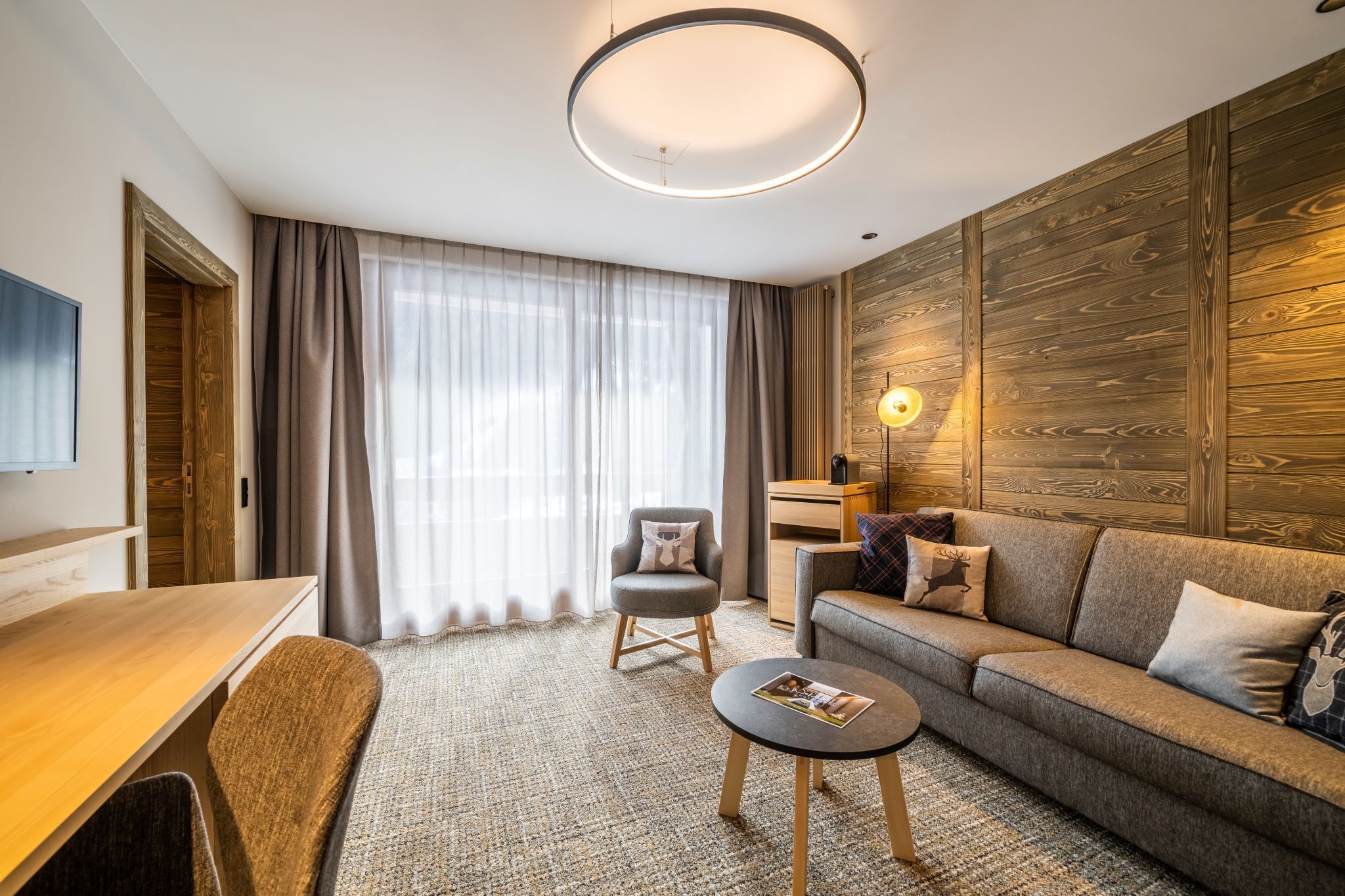 A modern hotel room with a wooden accent wall, a sofa, a chair, a small table, and a TV on the wall.