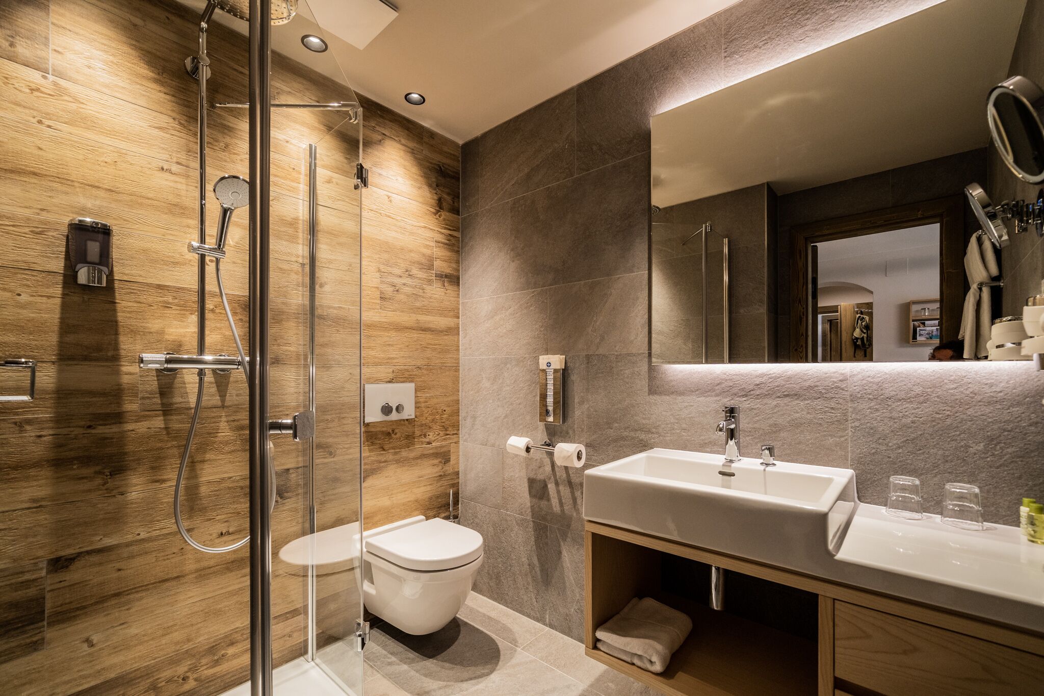 Un baño moderno con pared de madera, cabina de ducha de vidrio, inodoro blanco y un lavamanos grande con espejo.