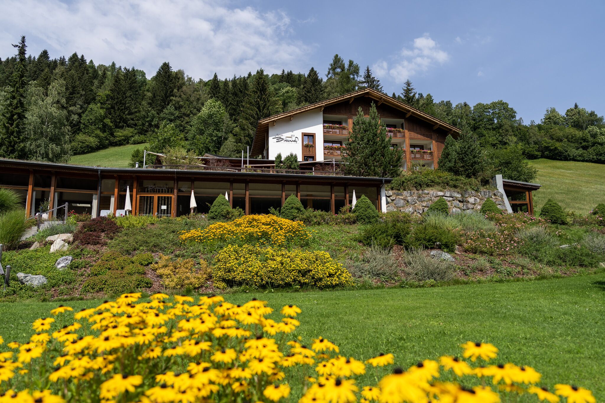 A two-story chalet with a balcony, surrounded by a lush garden and a forested hillside.