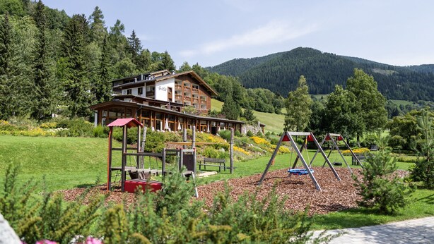 A lodge with a wooden exterior, a red-roofed play area, and a view of a forested mountain. - Hotel OTP Birkenhof (Bad Kleinkirchheim)