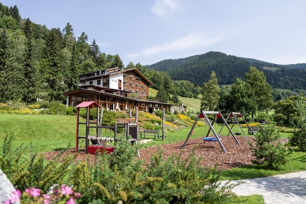 A lodge with a wooden exterior, a red-roofed play area, and a view of a forested mountain. - Hotel OTP Birkenhof (Bad Kleinkirchheim)