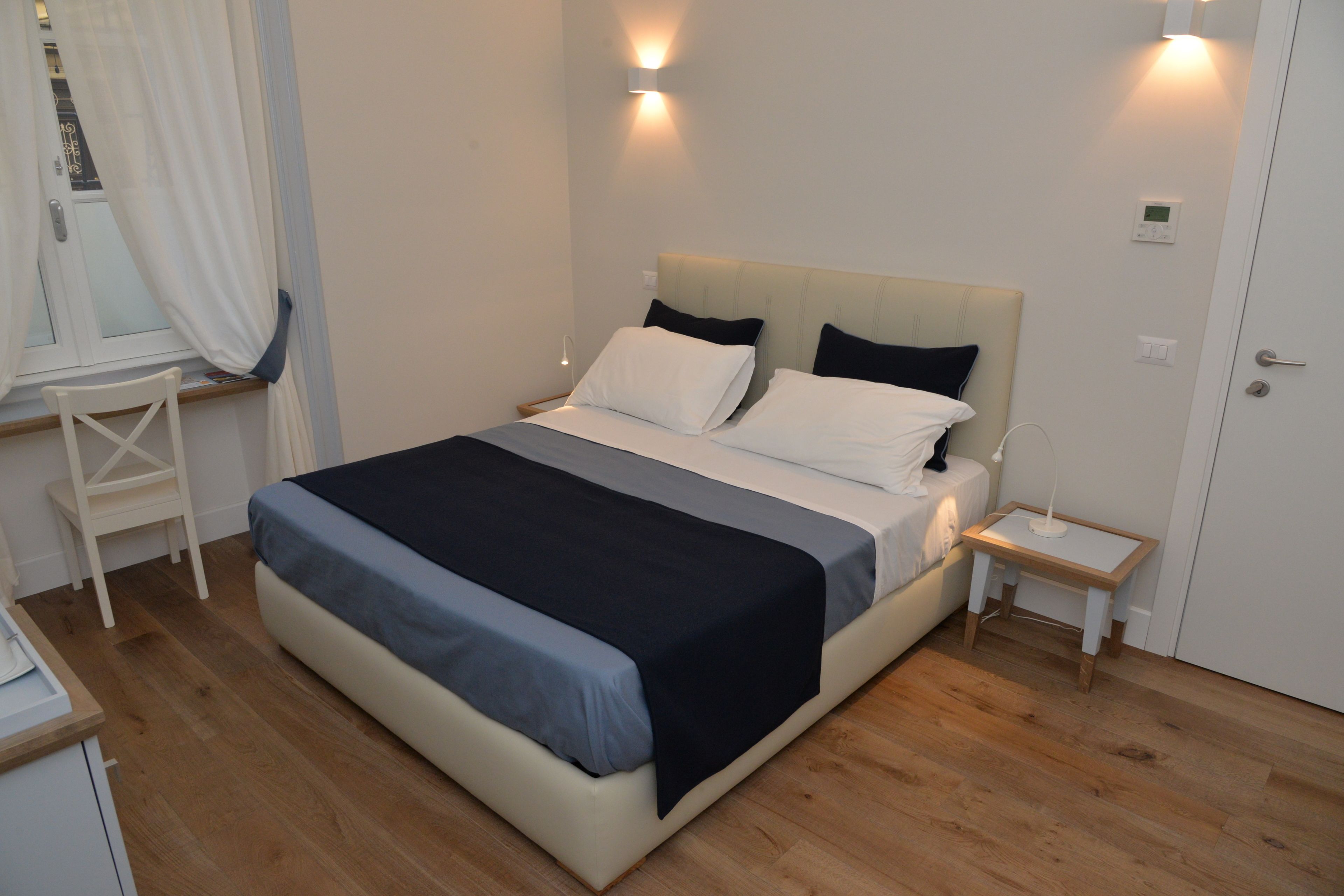Uma cama bem arrumada com um edredom cinza e branco, uma mesa de cabeceira com abajur e uma pequena cômoda de madeira.
