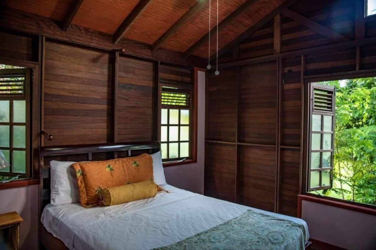 A wooden room with a bed, large windows, and a view of greenery outside.