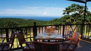 A wooden deck with a table set for dining, overlooking a scenic view of the ocean and hills. - Naniki Cottages (Surinam)