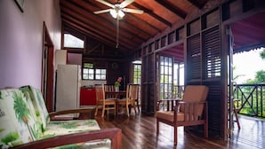A wooden-floored room with a ceiling fan, a dining table with chairs, a sofa, and a view of the outdoors through large windows. - Naniki Cottages (Surinam)