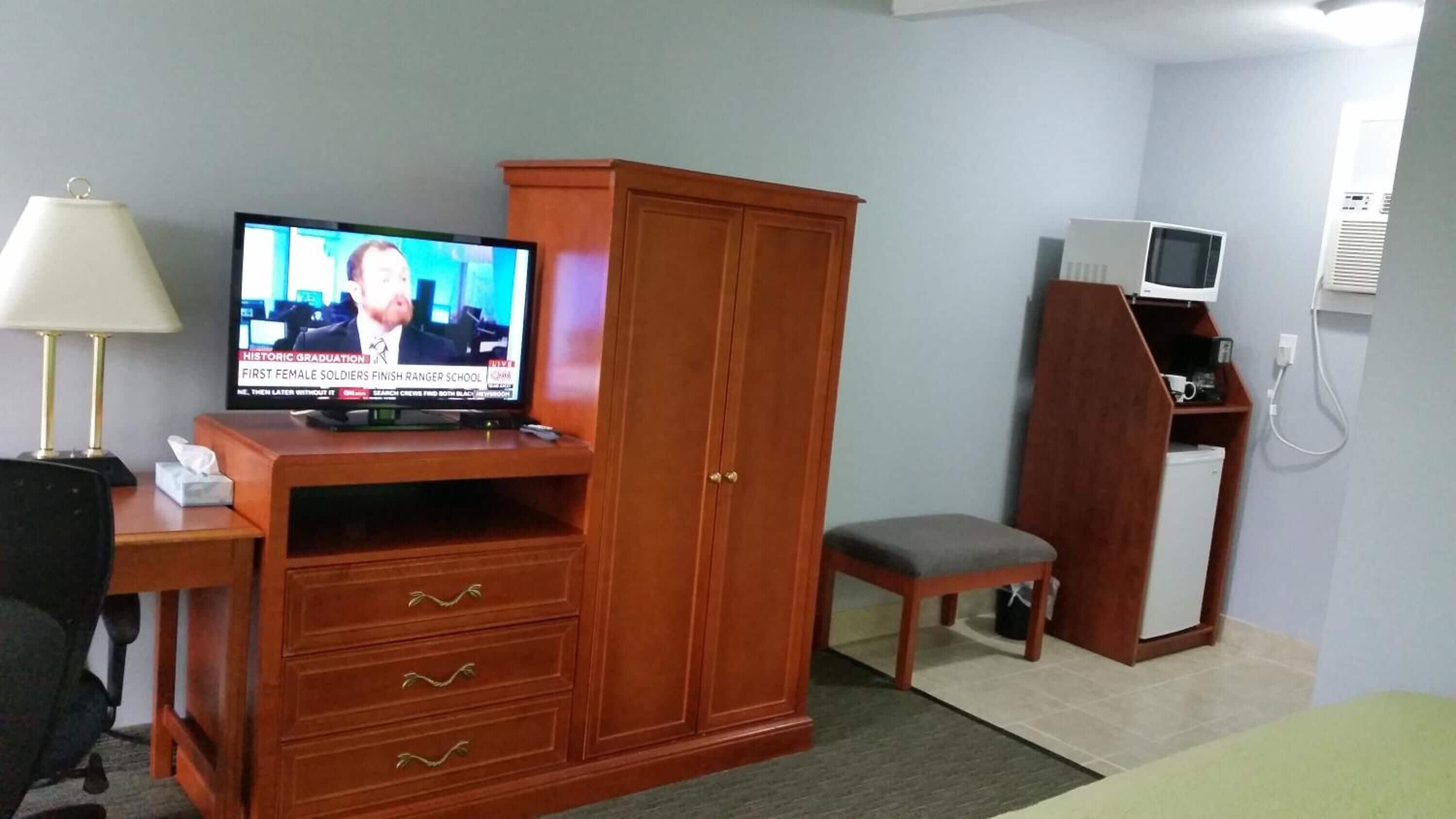 Habitación de hotel con un armario de madera, televisor, escritorio con lámpara, una silla y una pequeña zona de kitchenette.