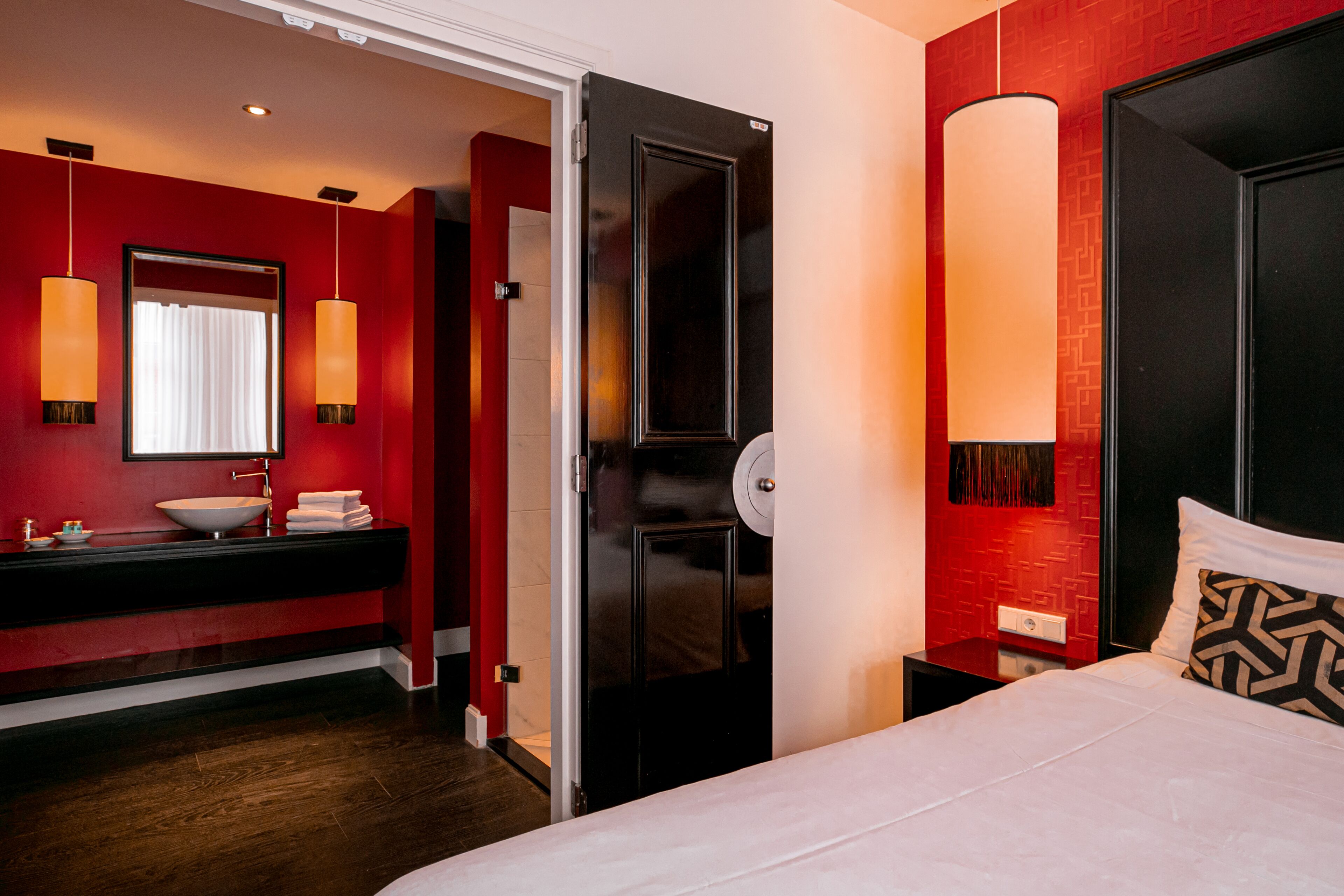 Een moderne hotelkamer met een rode accentmuur, een groot bed met een gekleurd kussen, een witte nachtkastje en een badkamer met een zwart wastafelmeubel en een witte wasbak.