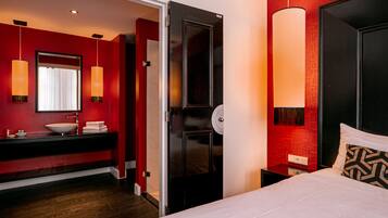 Een moderne hotelkamer met een rode accentmuur, een groot bed met een gekleurd kussen, een witte nachtkastje en een badkamer met een zwart wastafelmeubel en een witte wasbak.