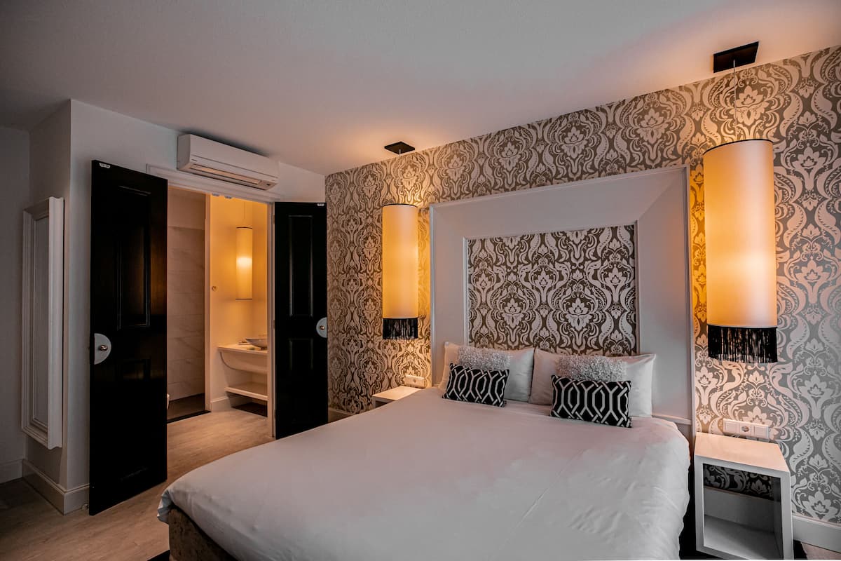 A bedroom with a large bed, two bedside tables, a nightstand, a wall-mounted lamp, and a bathroom door.