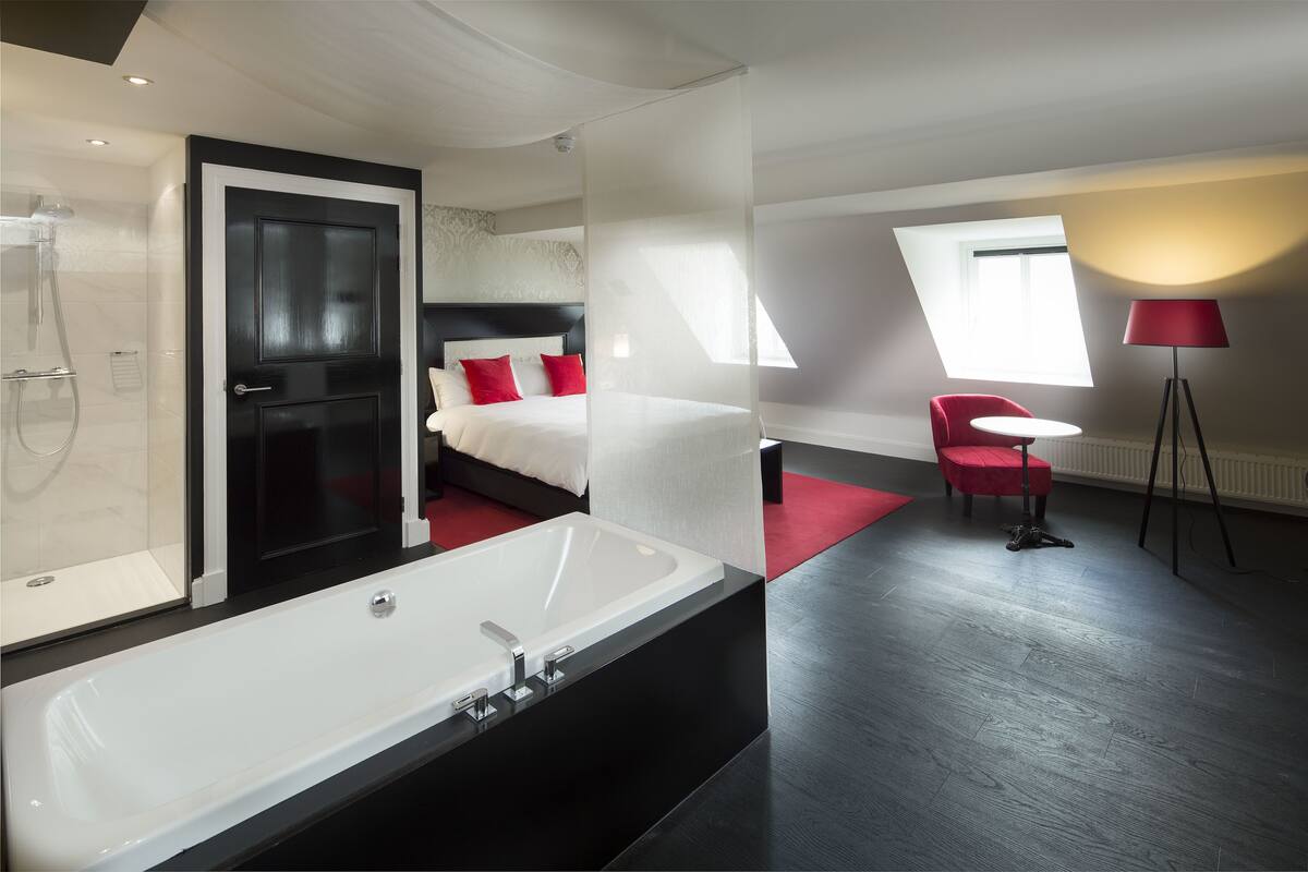 A modern hotel room with a large white bathtub, a separate shower area, a bed with red pillows, a red chair, and a floor lamp.