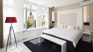 A modern hotel room with a large bed, a bench, a red floor lamp, and a view of a historic building. - Le Theatre Hotel (Maastricht)