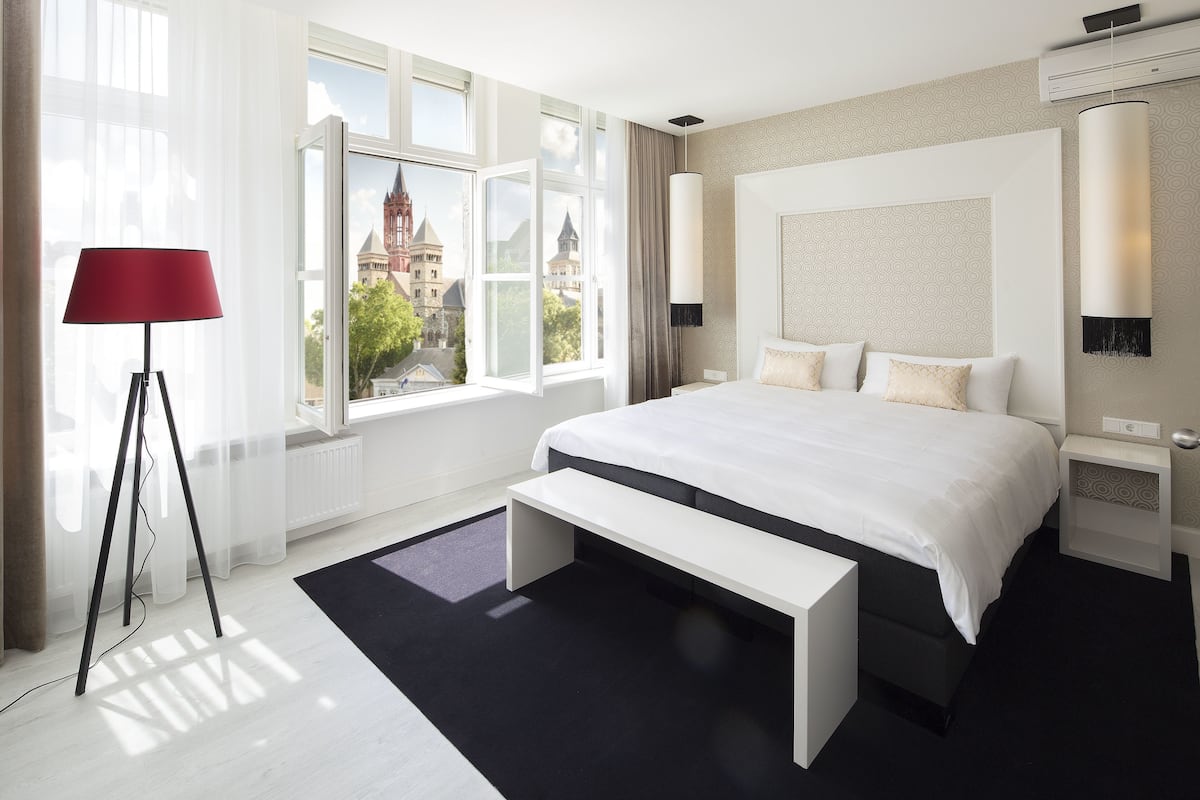 A modern hotel room with a large bed, a bench, a red floor lamp, and a view of a historic building.