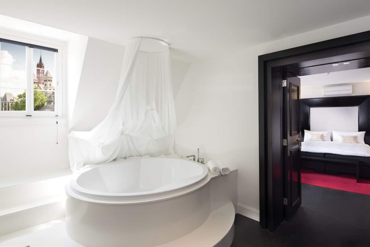 A modern bathroom with a freestanding bathtub, a window with a view of a building, and a separate bedroom with a bed and bedside table.