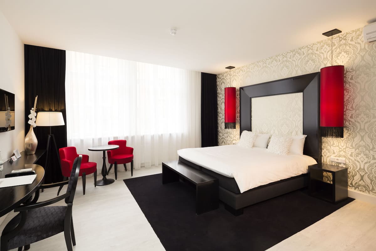 A modern hotel room with a large bed, a desk with a chair, a small table, and red chairs.