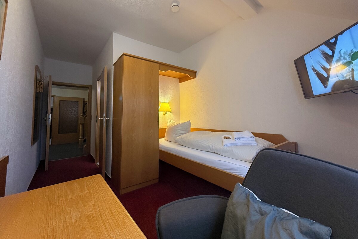 Ein kleines Hotelzimmer mit einem Einzelbett, einem Schreibtisch, einem Stuhl und einem an der Wand befestigten Fernseher.