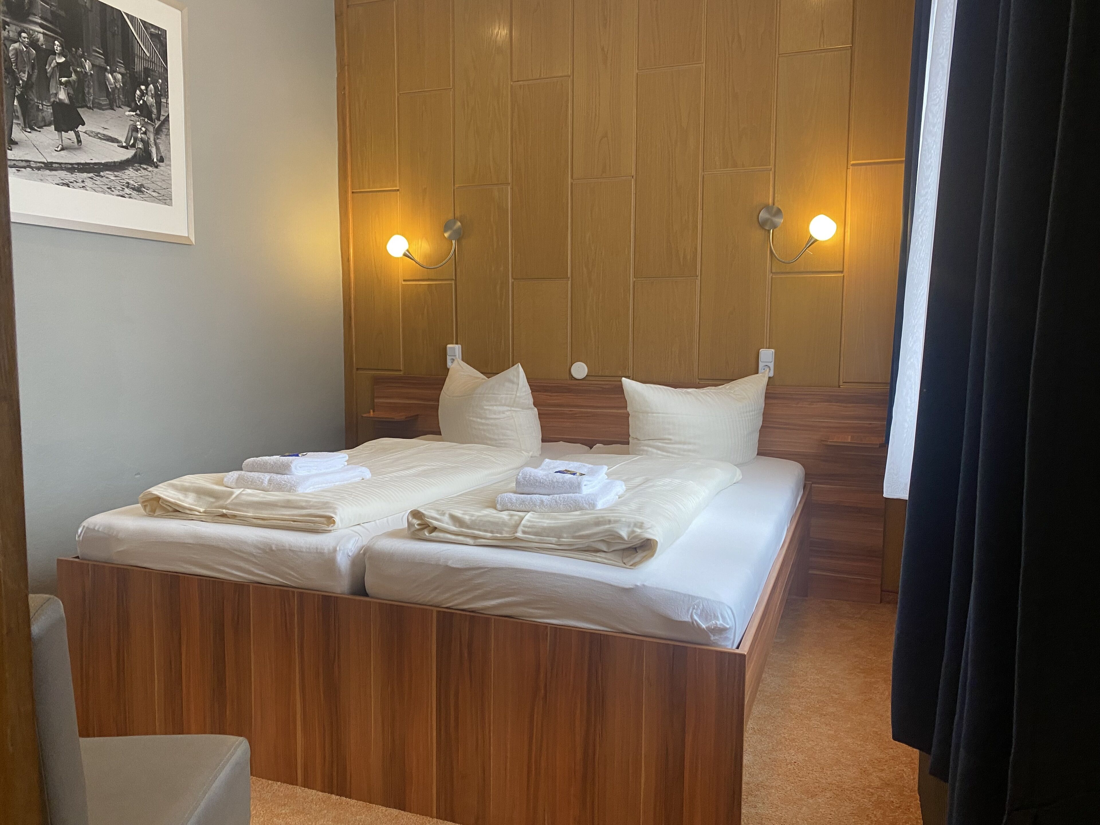 Habitación de hotel con cabecera de madera, dos lámparas de noche, cama con ropa blanca y toallas, y un cuadro en la pared.