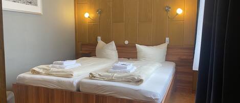 Habitación de hotel con cabecera de madera, dos lámparas de noche, cama con ropa blanca y toallas, y un cuadro en la pared.