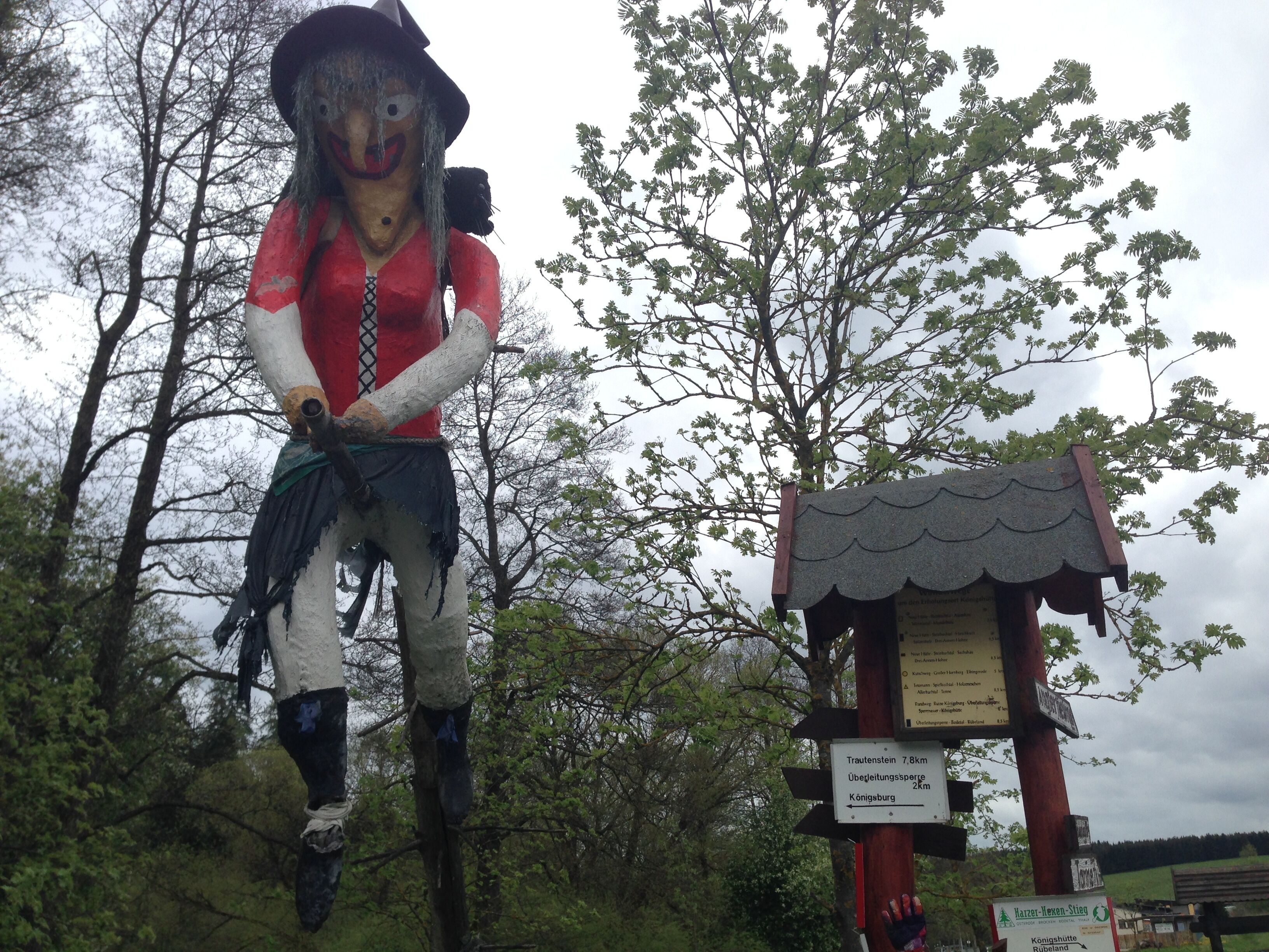 Une grande sculpture extérieure représentant un personnage vêtu d’un haut rouge et d’un pantalon noir, placée à côté d’un poteau indicateur en bois portant des informations sur les directions.