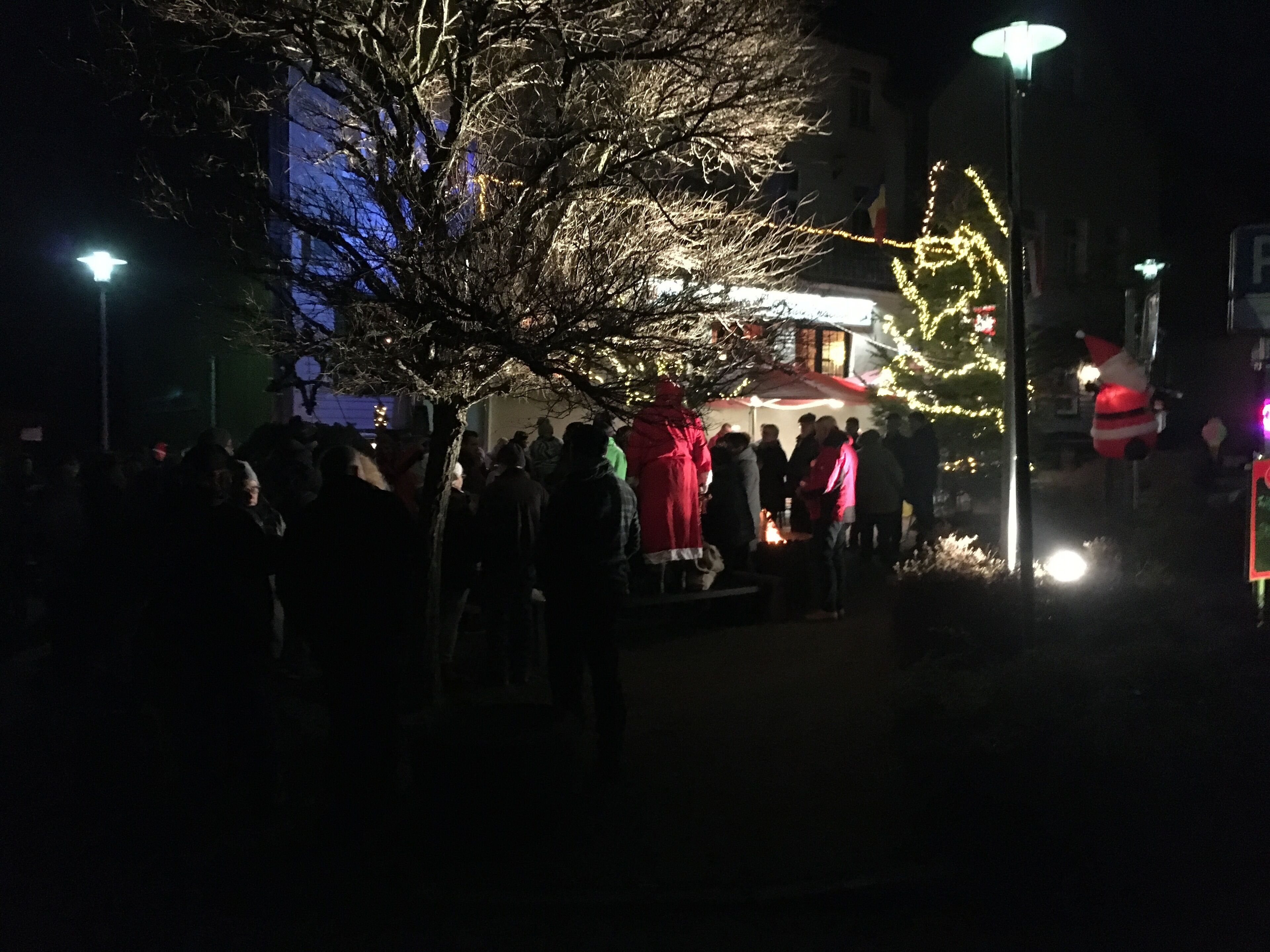 A nighttime outdoor gathering with people around a central fire, illuminated by street lamps and decorated trees.