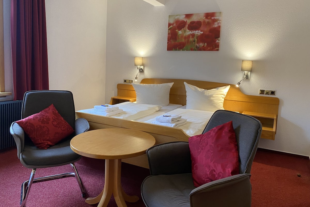 Habitación de hotel con una cama, dos mesitas de noche, dos lámparas, una mesa redonda, dos sillones y una cortina roja.