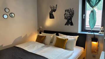 A neatly made bed with white linens and multiple pillows, a wall-mounted lamp, and a window with a curtain.