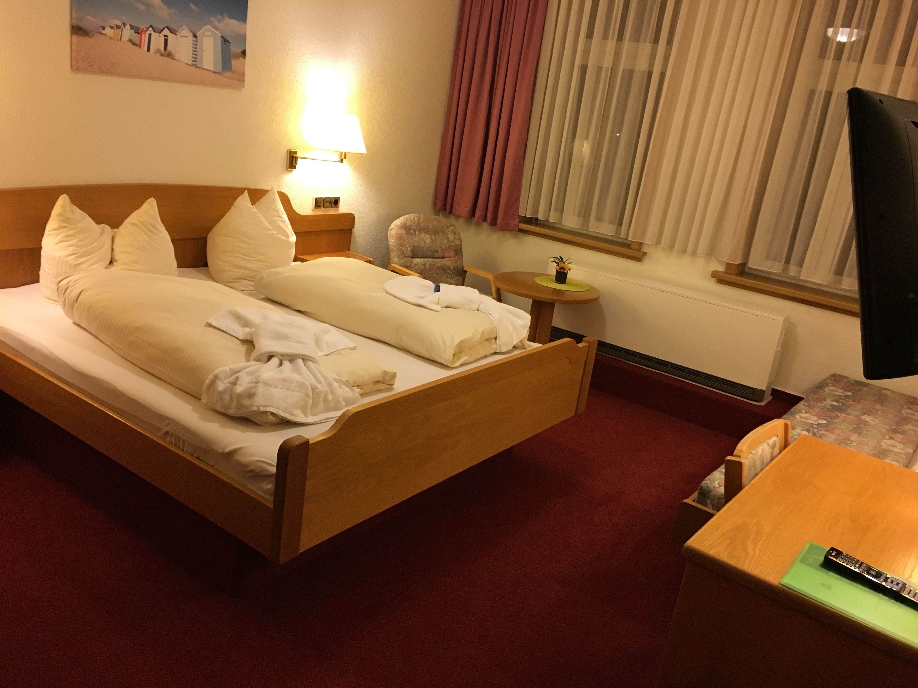 Ein Hotelzimmer mit einem hölzernen Bett, einem kleinen Tisch, einem Stuhl, einer Lampe und einem Fenster mit Vorhängen.